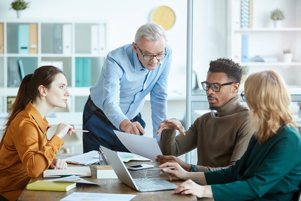 Diverse group of four business professionals in an office meeting, with an older male executive pointing to documents while a younger Black man in glasses discusses content next to two female colleagues.