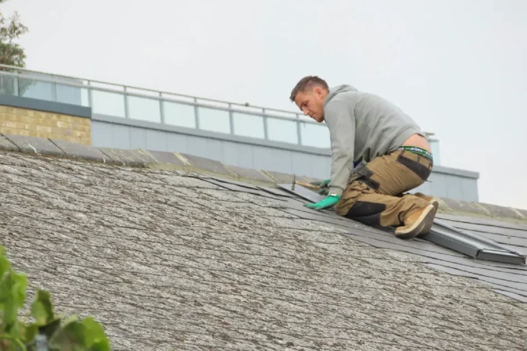 Roofer wearing a gray hoodie and khaki pants with green gloves kneeling on a mossy slate roof, performing roof repair or construction on a residential building.