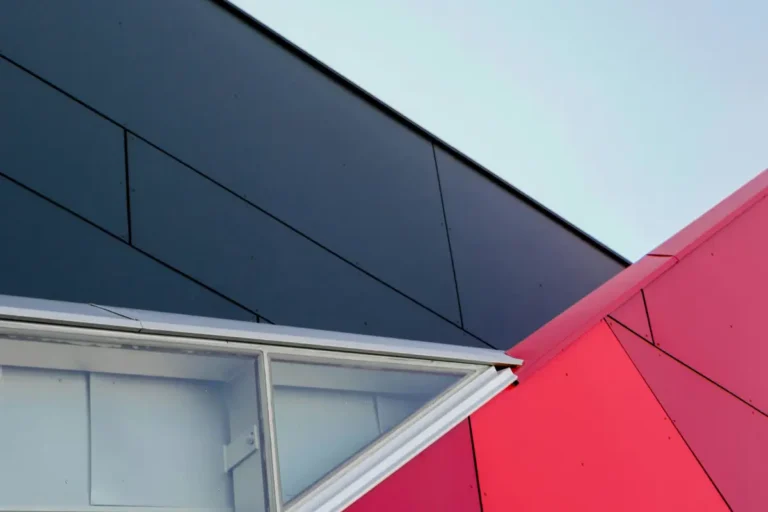 Abstract architectural close-up featuring the sharp, contrasting angles of black and red metal facade panels intersecting with a triangular glass skylight or window against a clear sky.