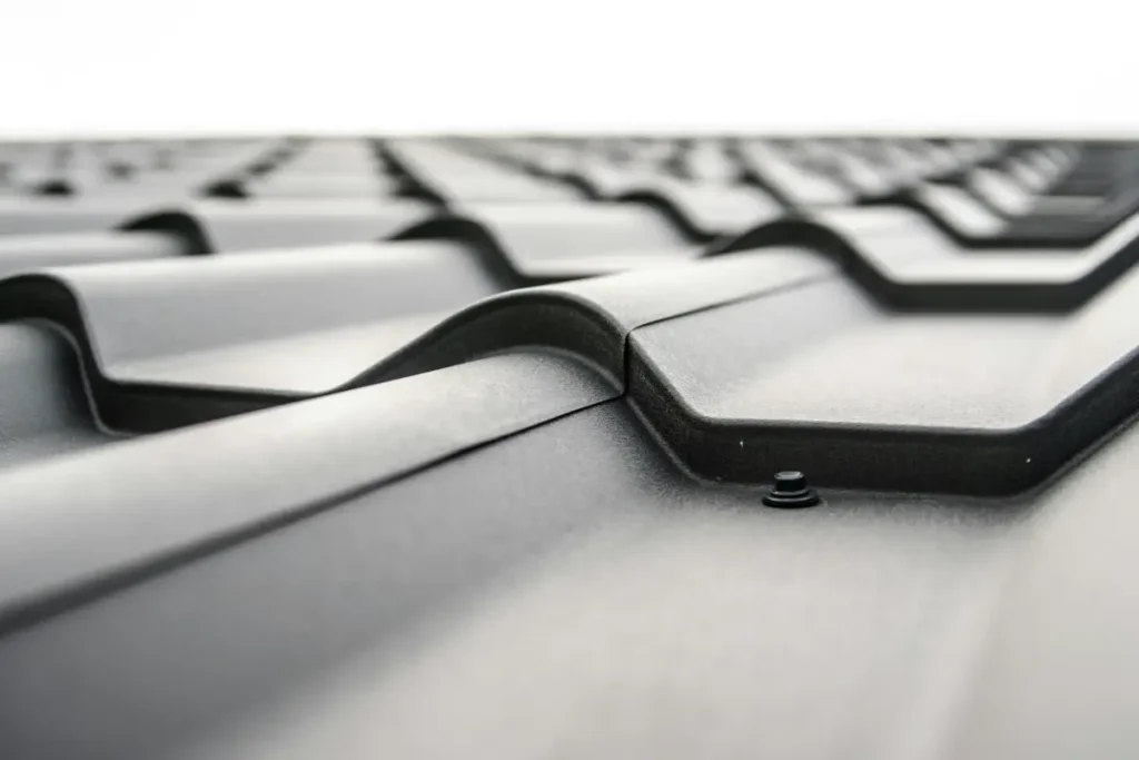 Close-up of black metal roofing tiles or sheets, showing their corrugated design and overlapping sections against a white or bright sky, with a roofing screw visible.