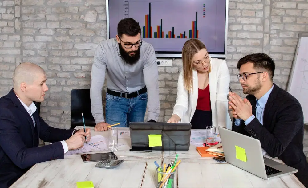 Digital Marketing team in a modern office discussing marketing strategies with charts and graphs on a screen in the background.