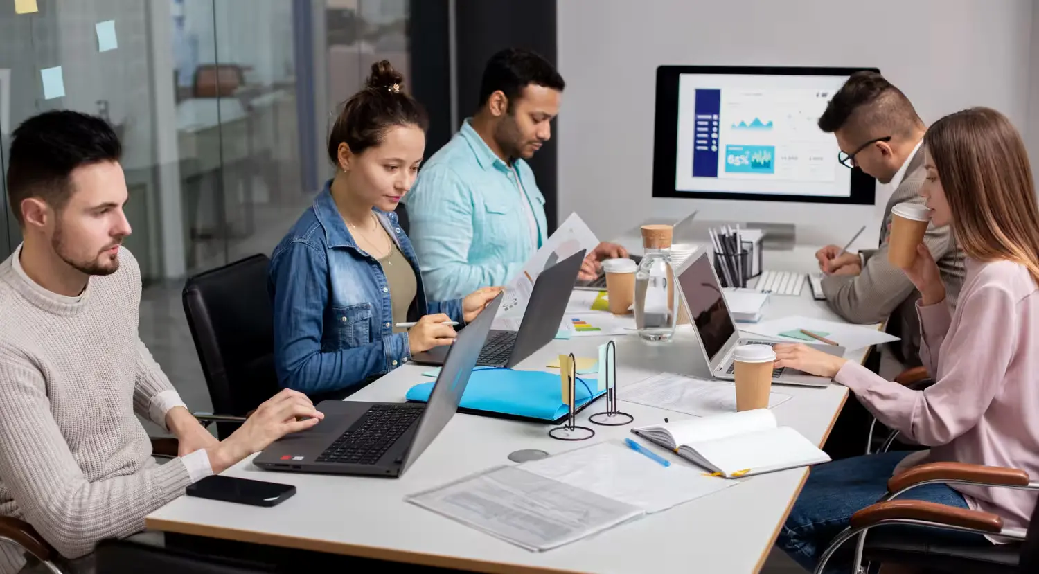 Diverse group of five professionals collaborating around a large table in an office, working on laptops, reviewing documents and data, and discussing a digital analytics report on a monitor.