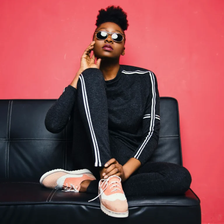 Stylish woman wearing a dark gray tracksuit with white stripes, round sunglasses, and two-tone pink and beige oxfords/sneakers while posing on a black leather couch against a bright pink background.